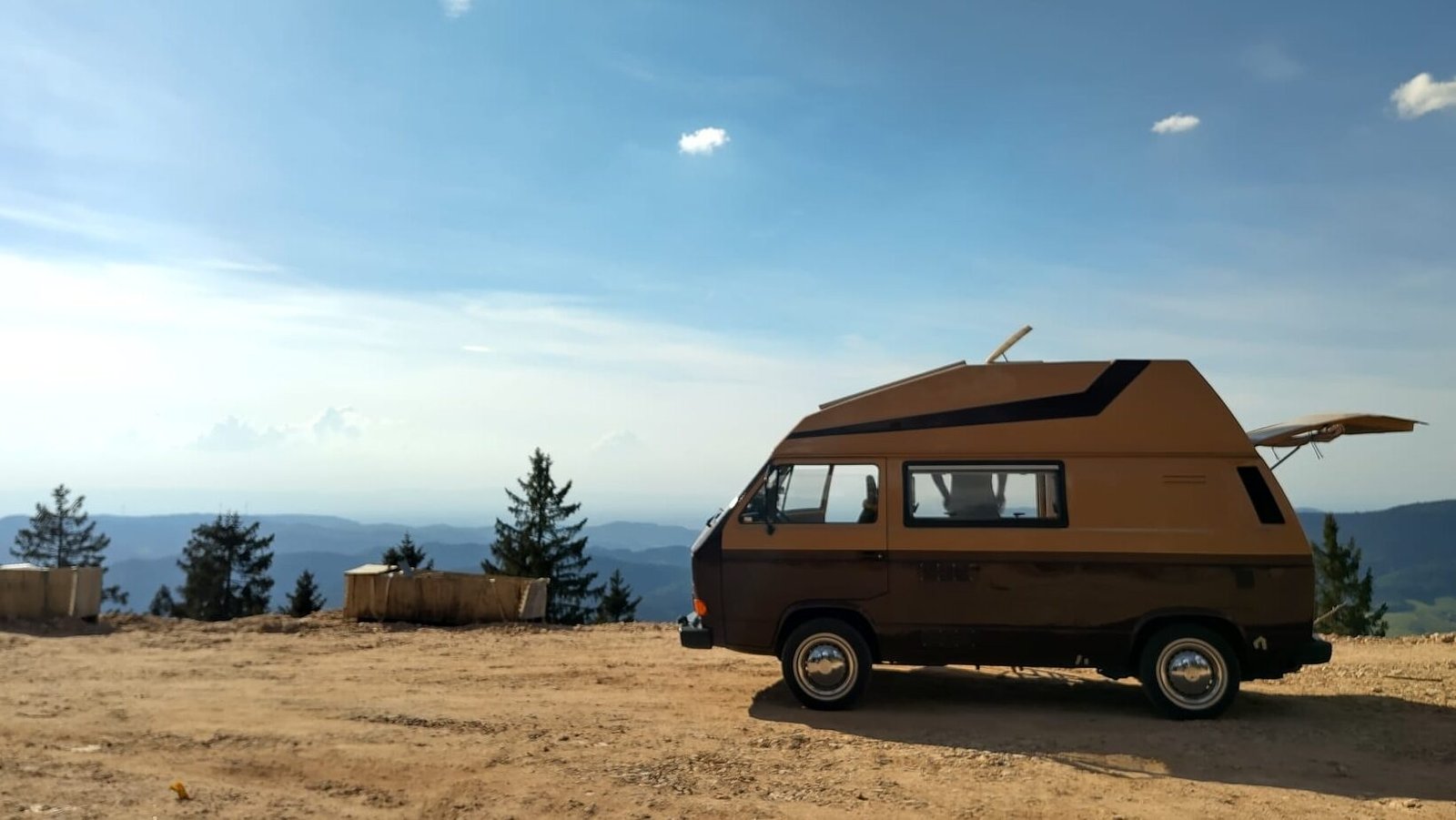Ein klassischer VW T3 Camper in zweifarbiger Braun-Beige-Lackierung steht mit geöffneter Heckklappe auf einem unbefestigten Plateau vor einer weiten Gebirgskulisse unter blauem Himmel.
